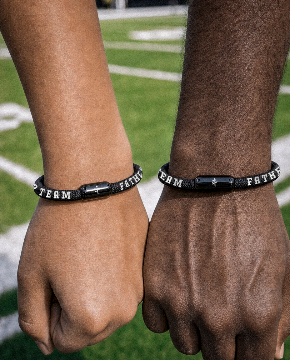 Two black braided bracelets with white lettering and a central black cross bead, worn on two different people's wrists with a blurred sports field in the background.